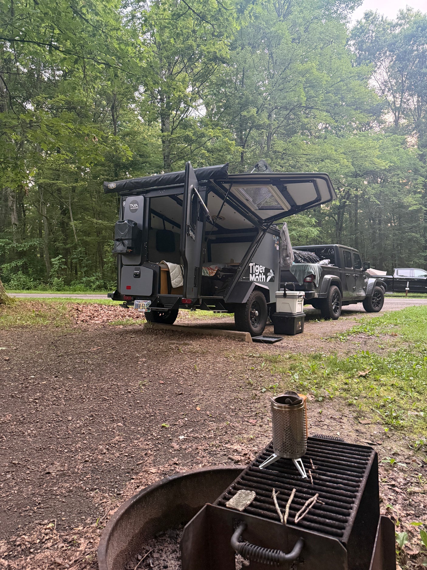 Explore the wild! This rv rental setup features a Tiger Moth overland trailer & Jeep at a forested campsite with a fire pit.