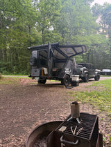 Explore the wild! This rv rental setup features a Tiger Moth overland trailer & Jeep at a forested campsite with a fire pit.