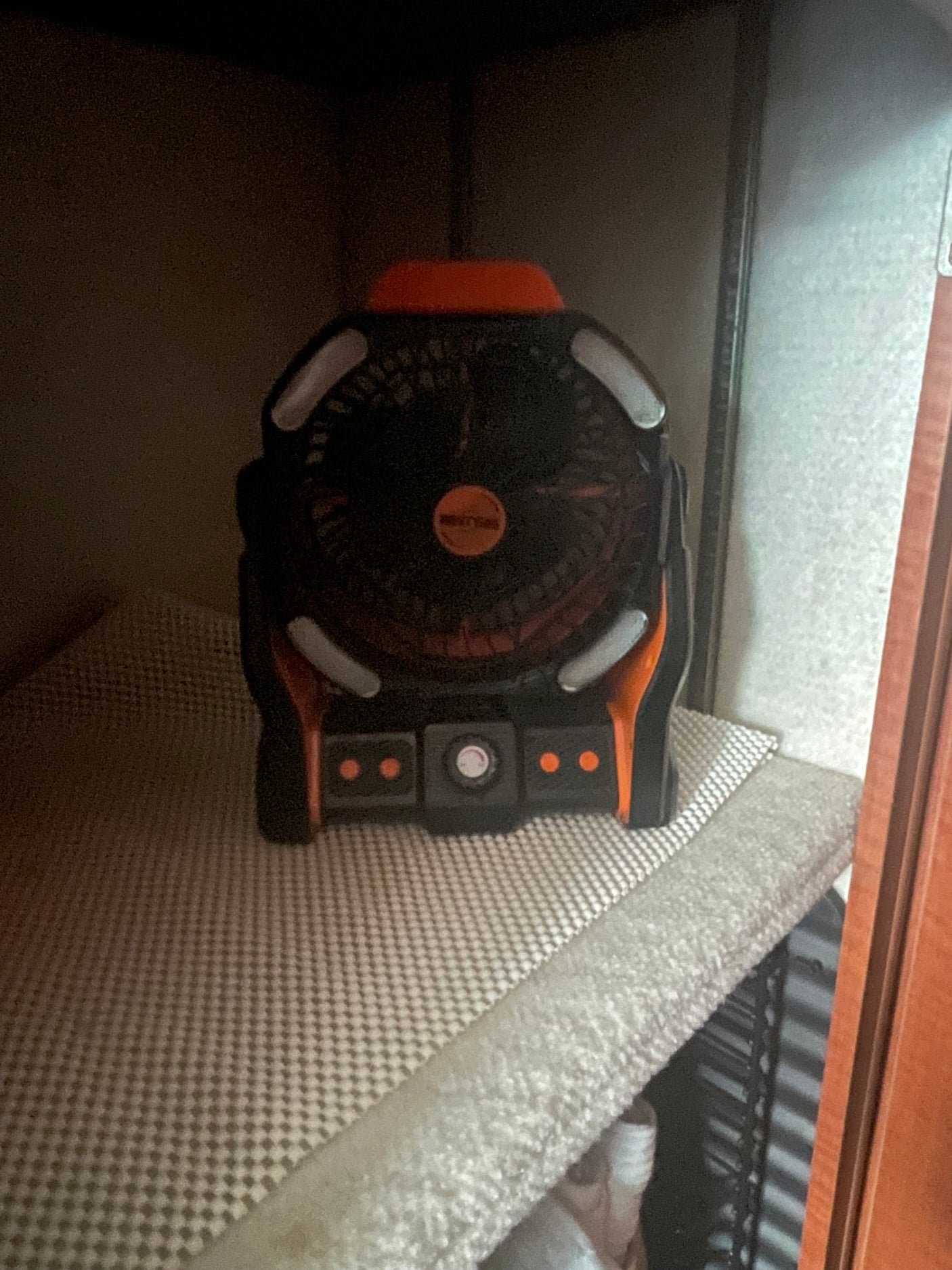 A black and orange portable fan, with a circular grill and front controls, rests on a textured shelf inside a dark RV rental cabinet.