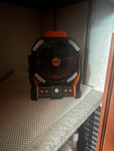 A black and orange portable fan, with a circular grill and front controls, rests on a textured shelf inside a dark RV rental cabinet.
