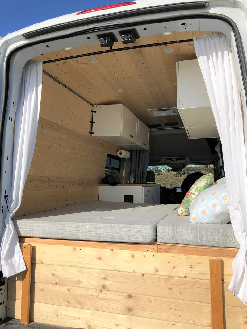  framed by white curtains. A wooden table holds a colorful snack board.|Experience tranquil van life in this RV rental: a cozy interior with wooden walls and seating — photo 14