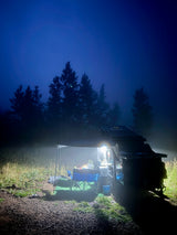 Illuminated rv rental provides a cozy camp setup with chairs & table under an awning in a misty, dark forest at night.