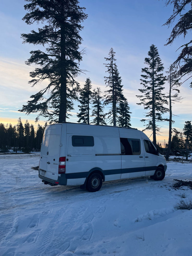  with beaming light rays and a white dog.|White camper van parked in snowy clearing with tall pines against a colorful sunset sky. Perfect winter RV rental escape.|White RV rental van at a mountain overlook. Snow-capped peaks glow under a blue sky with a crescent moon.|Explore this white RV rental camper van's interior: a compact mobile kitchen — photo 2