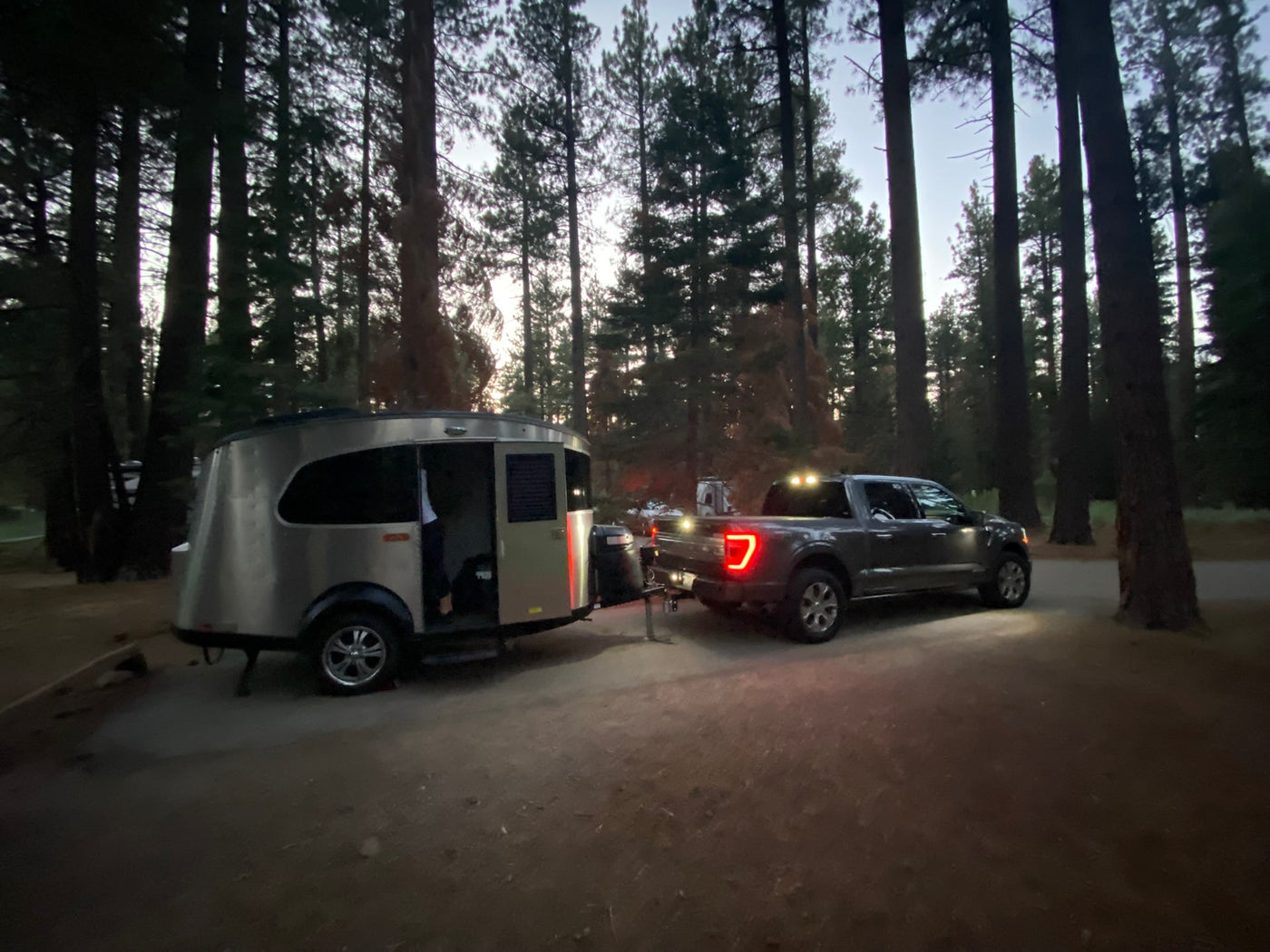 An RV rental trailer hitched to a pickup truck at a dim campsite surrounded by tall trees, ready for adventure. — photo 17