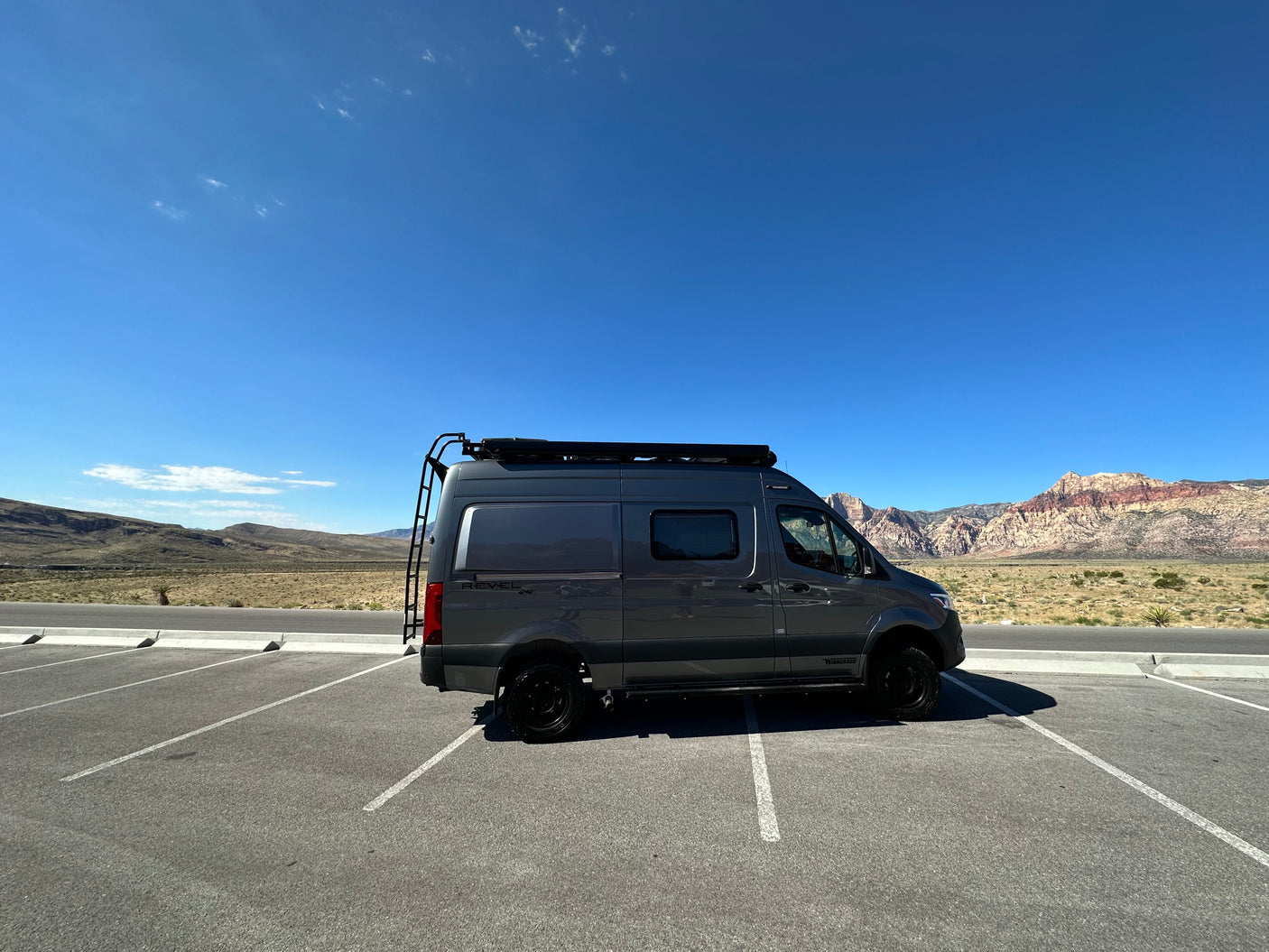 A dark gray Winnebago Revel 4x4 rv rental parked in a sunny desert landscape with mountains and a clear blue sky.