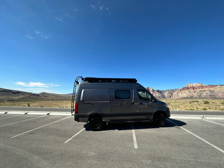 A dark gray Winnebago Revel 4x4 rv rental parked in a sunny desert landscape with mountains and a clear blue sky.