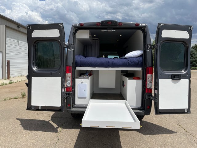 Experience comfort in this RV rental! Rear view of a dark gray camper van with open doors, revealing an elevated bed, interior storage, and pull-out tray.