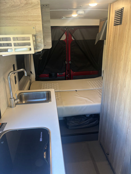 Compact rv rental interior showing kitchenette with sink/stove, bed, overhead cabinets, and rear privacy screens. Ideal for travel.