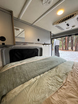 Cozy RV rental interior with bed, overhead storage, and snowy forest view through the open rear door.