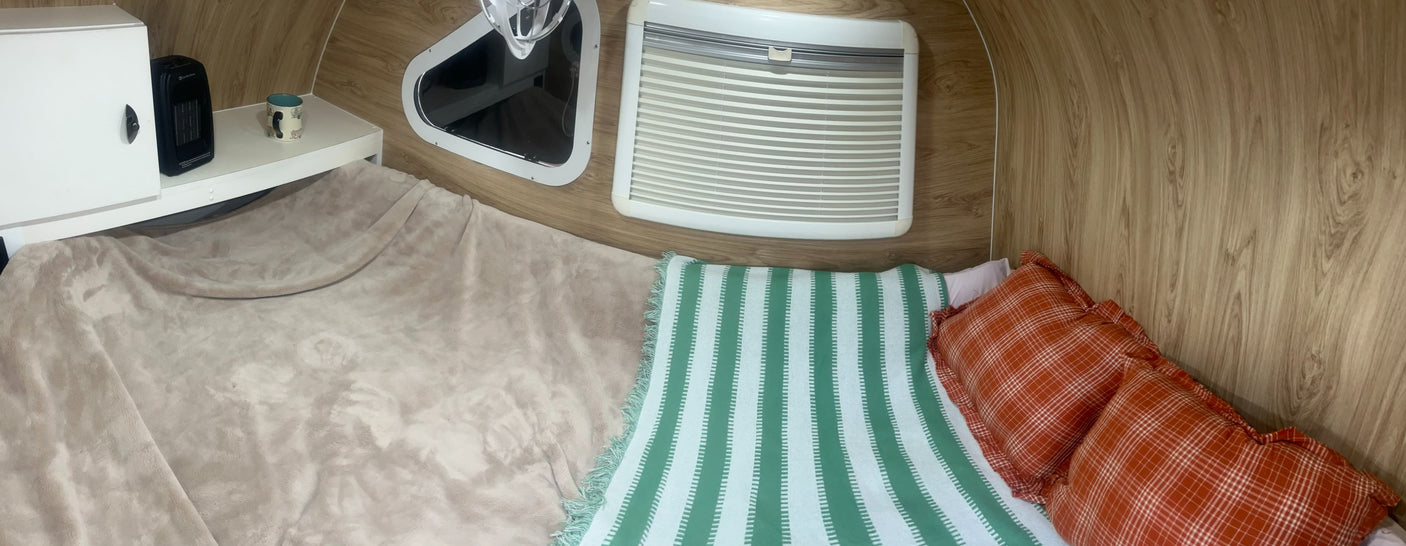 A compact RV rental interior features a bed with green striped and orange plaid bedding, a heater, and a window.