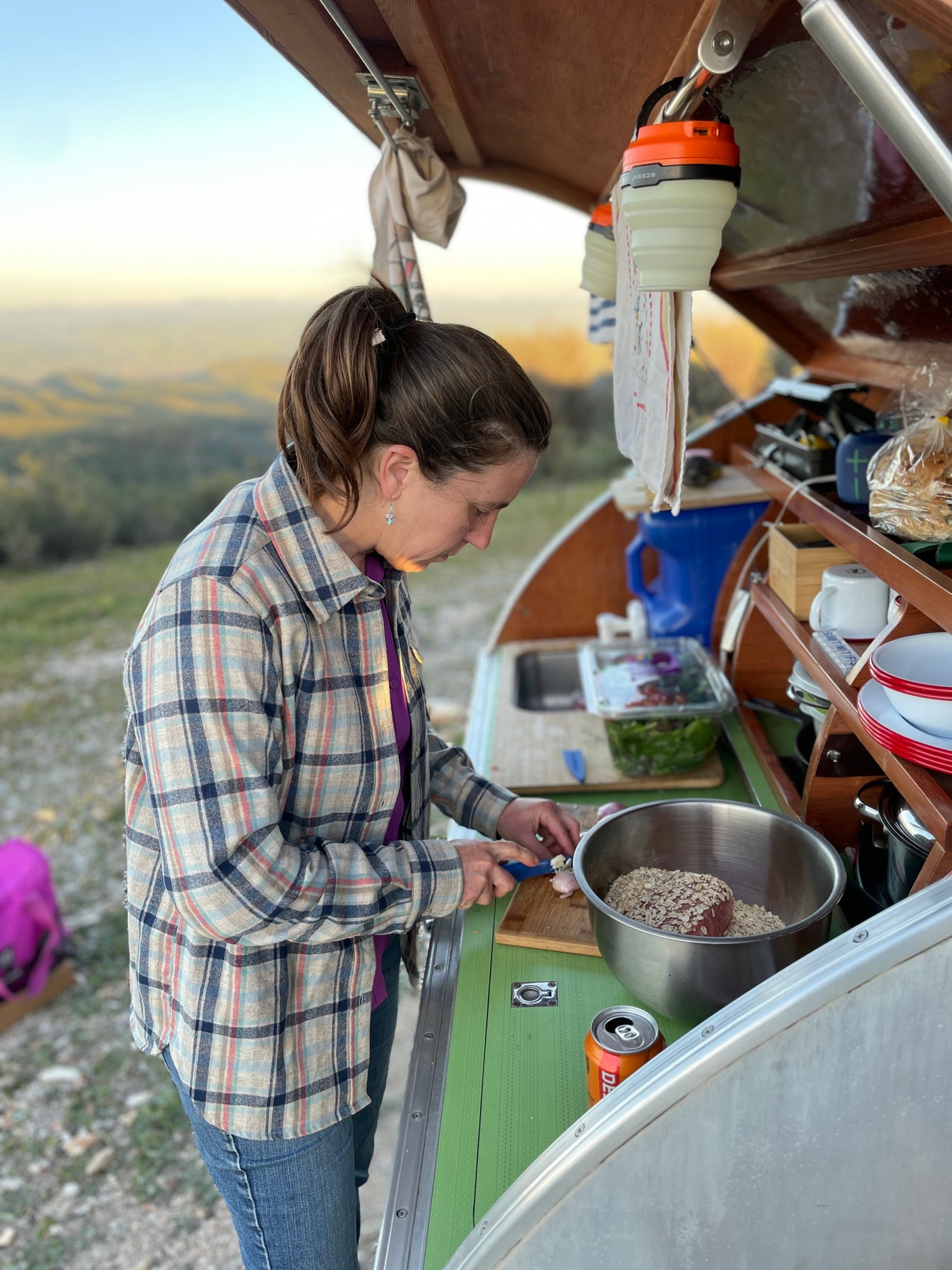 Woman chops ingredients at an RV rental's compact outdoor kitchen, with meat and oats in a bowl and a scenic view.