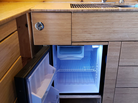 RV rental: Bamboo kitchen counter, sink, open illuminated mini-fridge, and cabinets with a round latch.