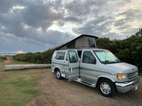 Silver pop-top RV rental parked by the ocean at sunset, ideal for beach camping adventures.