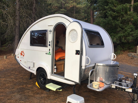 White teardrop **RV rental** parked in a forest campsite. Its door is open, showing a cozy interior and exterior setup.