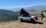 Adventure awaits! This white SUV with a rooftop tent, available for RV rental, offers elevated camping in the mountains with stunning valley views.