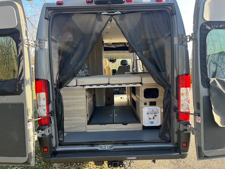 Inside this rv rental camper van, open rear doors show a comfortable bed, lower storage, and water hookup, with a screen.