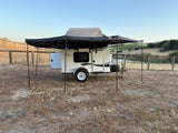 RV rental: Small camper trailer with a large awning and rooftop tent, parked in a scenic outdoor setting.