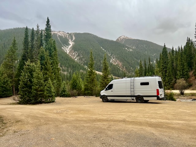  and 3 dogs relax on a blue mat in the sand.|Explore the desert in comfort with an RV rental. A white camper van with an awning faces arid mountains under a blue sky.|White rv rental camper van parked on dirt amidst a lush evergreen forest & mountains under an overcast sky.|White RV rental van parked in a scenic mountain lot with snow-dusted peaks and lush trees. Ready for travel!|White RV rental van parked alone in a scenic mountain lot. Blue sky & green trees enhance the travel vibe.|White camper van par — photo 3