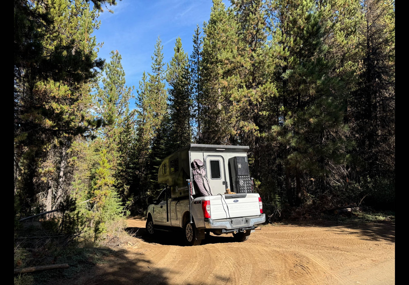Experience nature with an RV rental: a robust truck camper parked on a dirt road among tall pine trees, ready for adventure.