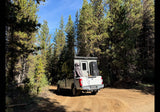 Experience nature with an RV rental: a robust truck camper parked on a dirt road among tall pine trees, ready for adventure.