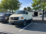 Weathered white commercial van, similar to one for RV rental, parked in a sunlit lot with green trees and buildings.