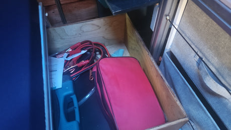 RV rental utility storage: Wooden drawer contains jumper cables, a vehicle charger, and a red emergency kit bag.