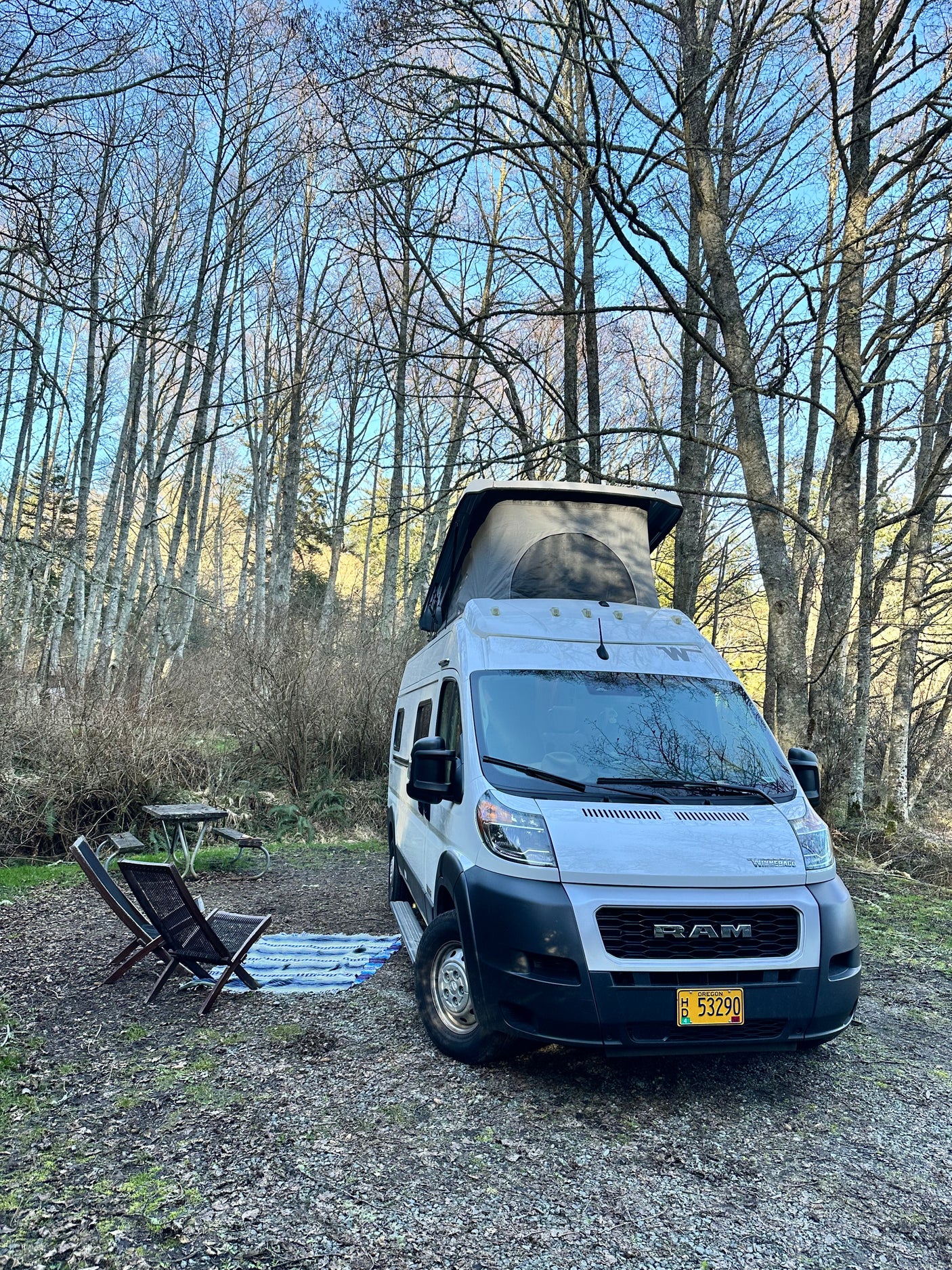 White pop-top RV rental camper van at a wooded campsite with chairs & picnic table for a peaceful getaway.