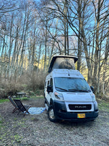 White pop-top RV rental camper van at a wooded campsite with chairs & picnic table for a peaceful getaway.