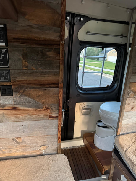 Explore this RV rental! Rustic wood interior, control panel, portable toilet, and cozy cushioned seating in a functional camper van setup.