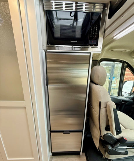 RV rental interior shows a compact stainless steel microwave over a refrigerator, next to a comfy driver's seat.