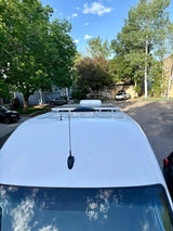 White RV rental van's roof: solar panels, antenna, and a sunny residential backdrop with trees.