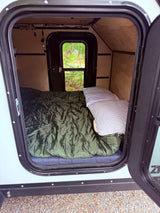 A cozy RV rental interior features a bed with green bedding and white pillows. A window reveals lush green trees outside.