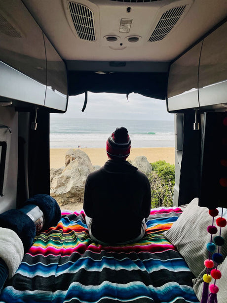 RV rental escape: Person enjoys tranquil beach & ocean view from inside a camper van, resting on a striped blanket.