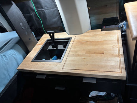 RV rental kitchen: black sink, wooden countertop with cutting board, and nearby bed in a compact van interior.