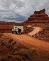 White RV rental van on a winding red dirt road in a desert landscape with large rock formations under a cloudy sky.