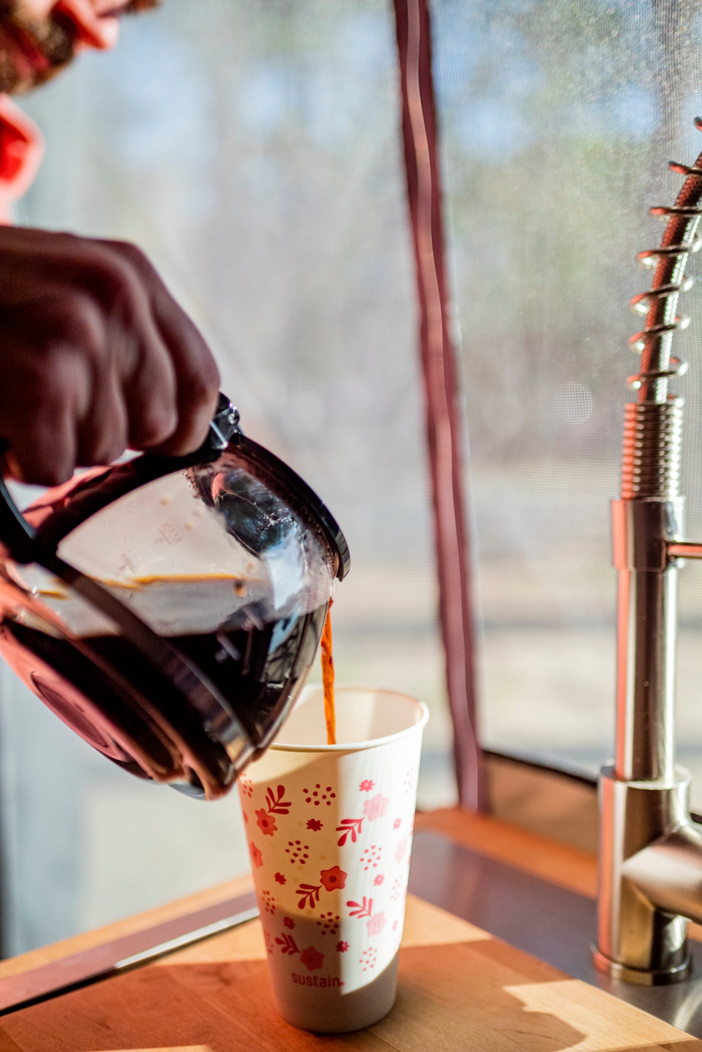 A hand pours coffee into a 'sustain' paper cup from a carafe, showing an RV rental kitchen setup with a faucet.