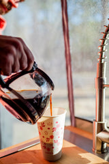 A hand pours coffee into a 'sustain' paper cup from a carafe, showing an RV rental kitchen setup with a faucet.