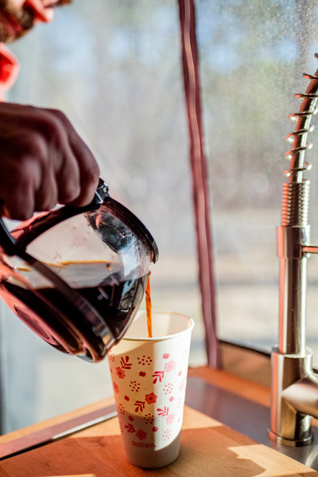 A hand pours coffee into a 'sustain' paper cup from a carafe, showing an RV rental kitchen setup with a faucet.