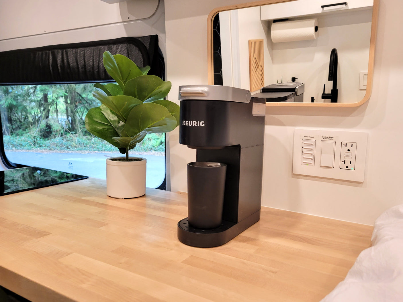 Compact rv rental kitchen area with a Keurig coffee maker, plant, wood countertop, and window overlooking nature.