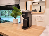 Compact rv rental kitchen area with a Keurig coffee maker, plant, wood countertop, and window overlooking nature.