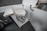 RV rental interior showcasing a compact, white composting toilet with open cabinet doors on a dark, textured floor.