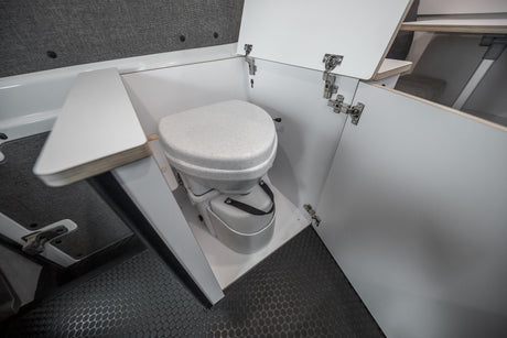 RV rental interior showcasing a compact, white composting toilet with open cabinet doors on a dark, textured floor.