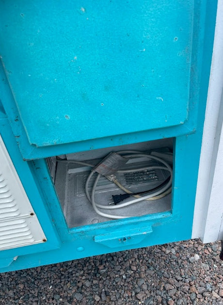 Open electrical compartment on an **rv rental**, revealing power cables and a plug. Teal exterior, gravel below.