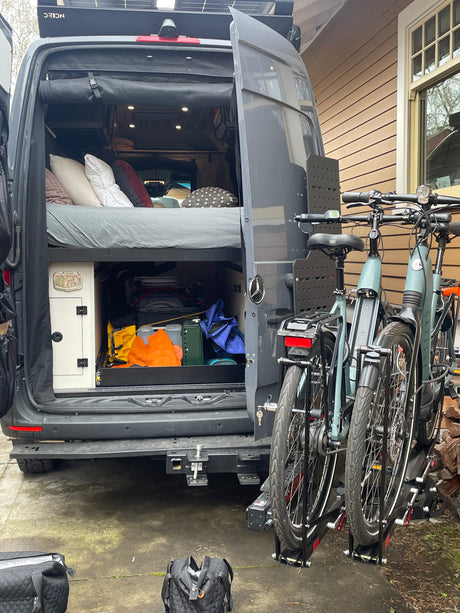 Dark grey camper van with open rear doors showing bed and storage. Roof solar and two e-bikes on rack. Perfect RV rental for adventure!