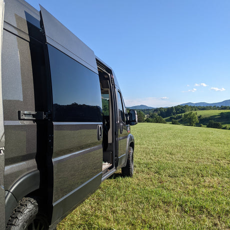 Dark camper van (RV rental) with its side door open, parked on a lush green hillside overlooking distant mountains.