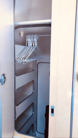 Open closet in an RV rental, with a metal hanging rod holding white wire hangers. It includes left-side shelves and a dark hose above.