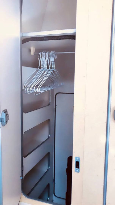 Open closet in an RV rental, with a metal hanging rod holding white wire hangers. It includes left-side shelves and a dark hose above.