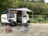 Relaxing lakeside with an RV rental setup: white camper, awning, chairs, and a cozy rug by the pond.