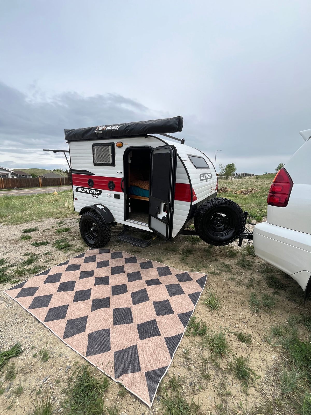 An RV rental: A small white & red teardrop camper hitched to an SUV, door open to a bed, next to an outdoor mat. — photo 8
