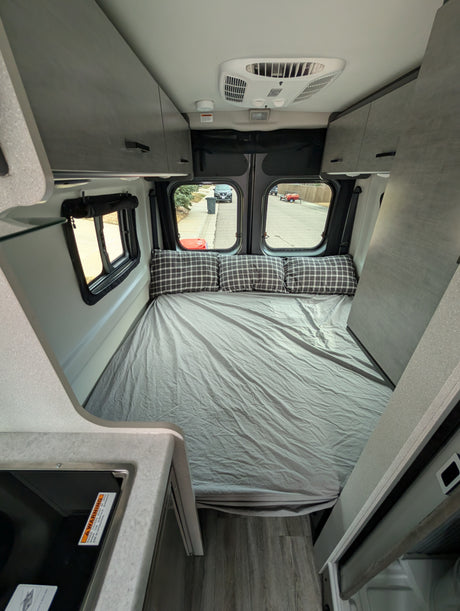 Comfortable RV rental interior with a large bed, plaid pillows, kitchen counter, and overhead storage, ideal for compact travel.